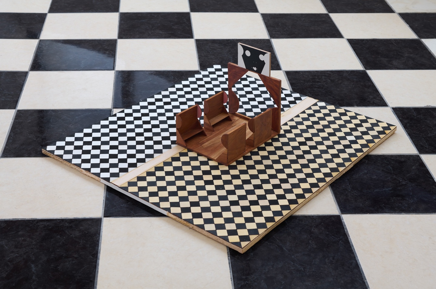 Atop a black-and-white checkboard floor sits a smaller, black-and-white checkerboard piece, at an angle; a brown seating structure with four chair-like features plus a white panel with a black face is on top of that.