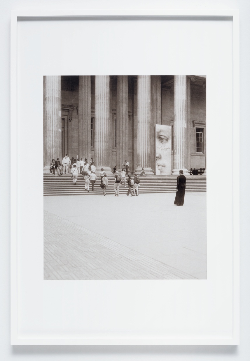 A black-and-white photograph shows a crowd of people walking up the steps of a large classical-style building while a figure in a long modest black shift stands watching the crowd.