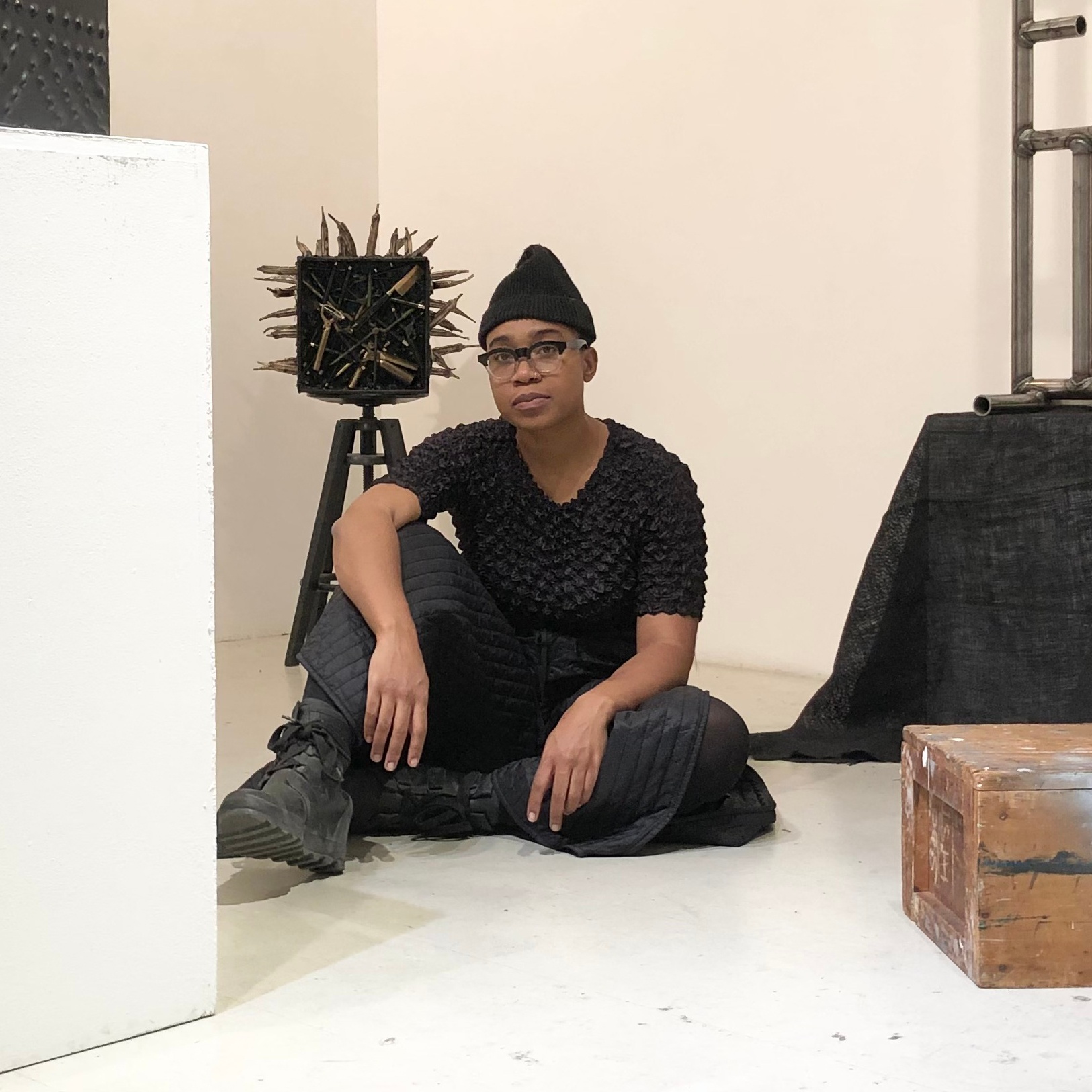 A Black artist sits casually on the floor of a white-walled gallery space, with one arm draped over her leg. She looks directly at us, dressed in all black wearing cap, glasses, t-shirt, pants, and boots. Around her and sculptures sitting on the ground and on a pedestal. One sculpture behind her shoulder includes spiky protrusions from a square surface. 