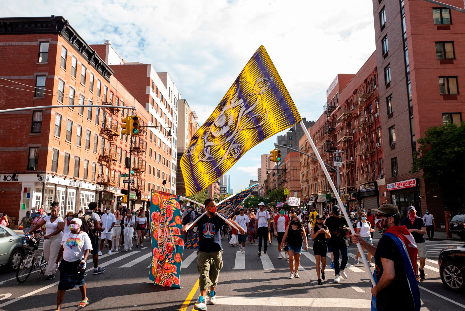 A photograph of a crowd walking in the street. A Black man in the center carries two flags, and a person in the foreground waves a purple and yellow flag.