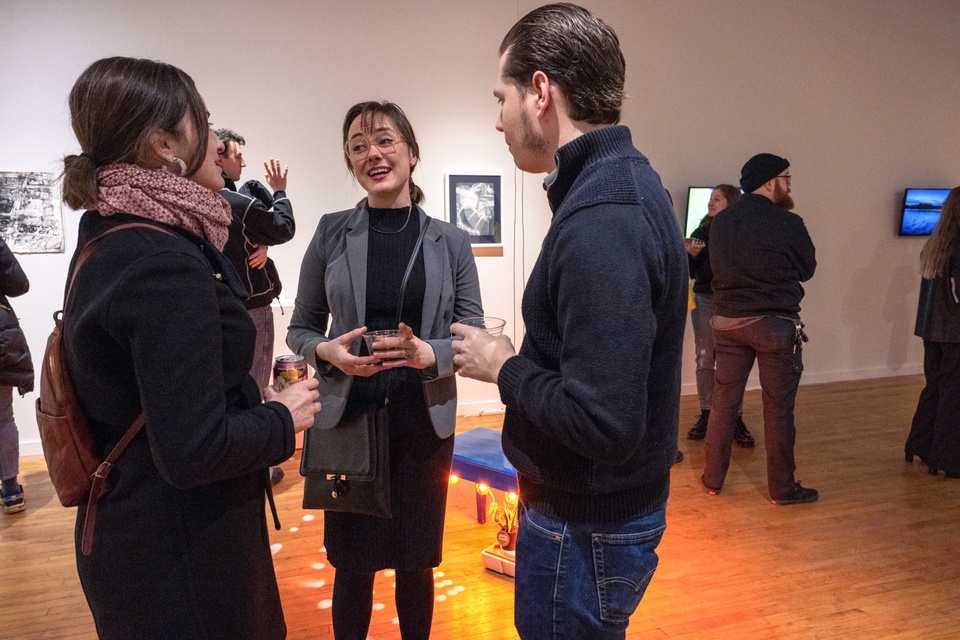 Three people stand and chat in a gallery space. The floor behind them is lit orange by a sculpture sitting on the floor.