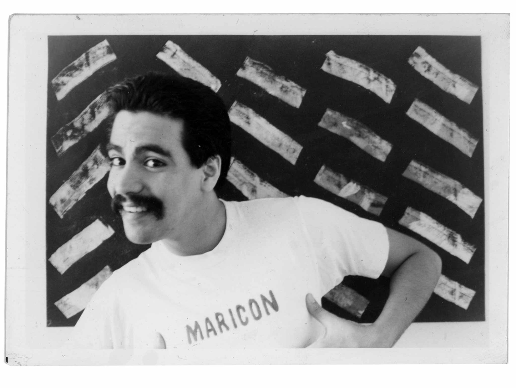 A black and white photo of a Latino man smiling standing and wearing a shirt that says "MARICON."