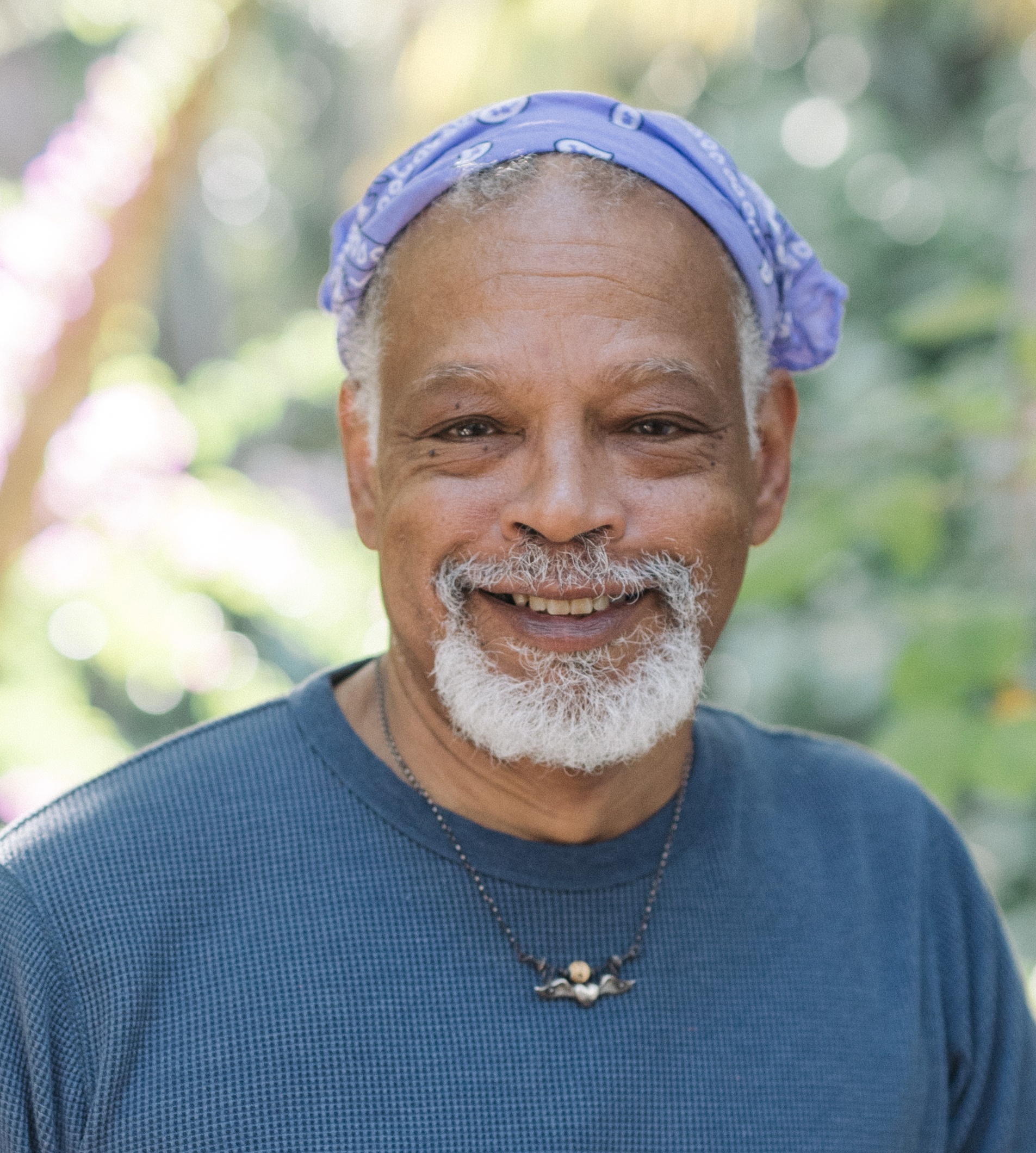 A headshot of Ishmael Houston-Jones, a man with light brown skin and a gray mustache and goatee. He smiles broadly against a background of trees and leaves. He wears a lilac bandana around the crown of his head and a blue shirt. Photo by Mark Poucher