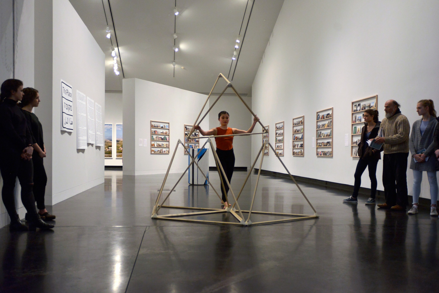 A crowd watches as a young, Asian, female dancer poses with a wooden triangular sculpture.