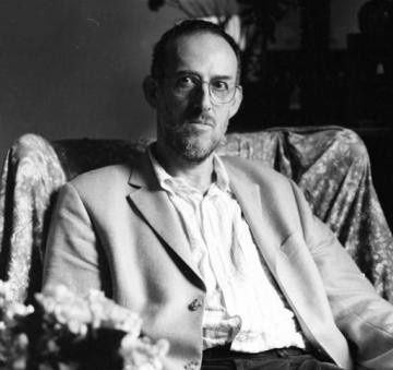 A man in glasses and a casual blazer sits in an armchair. 