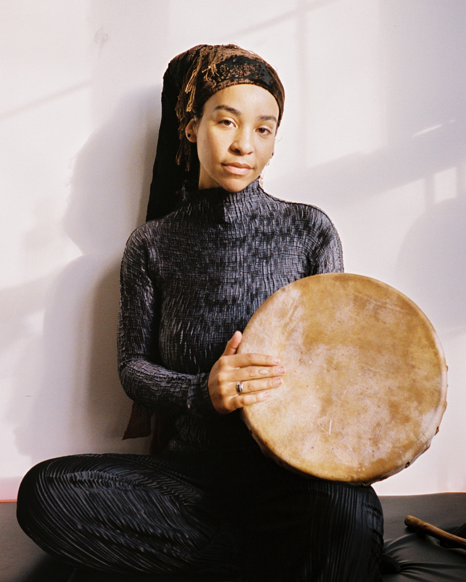 A photo of the artist AnAkA. She sits in light and shadow from a window while holding a drum. 