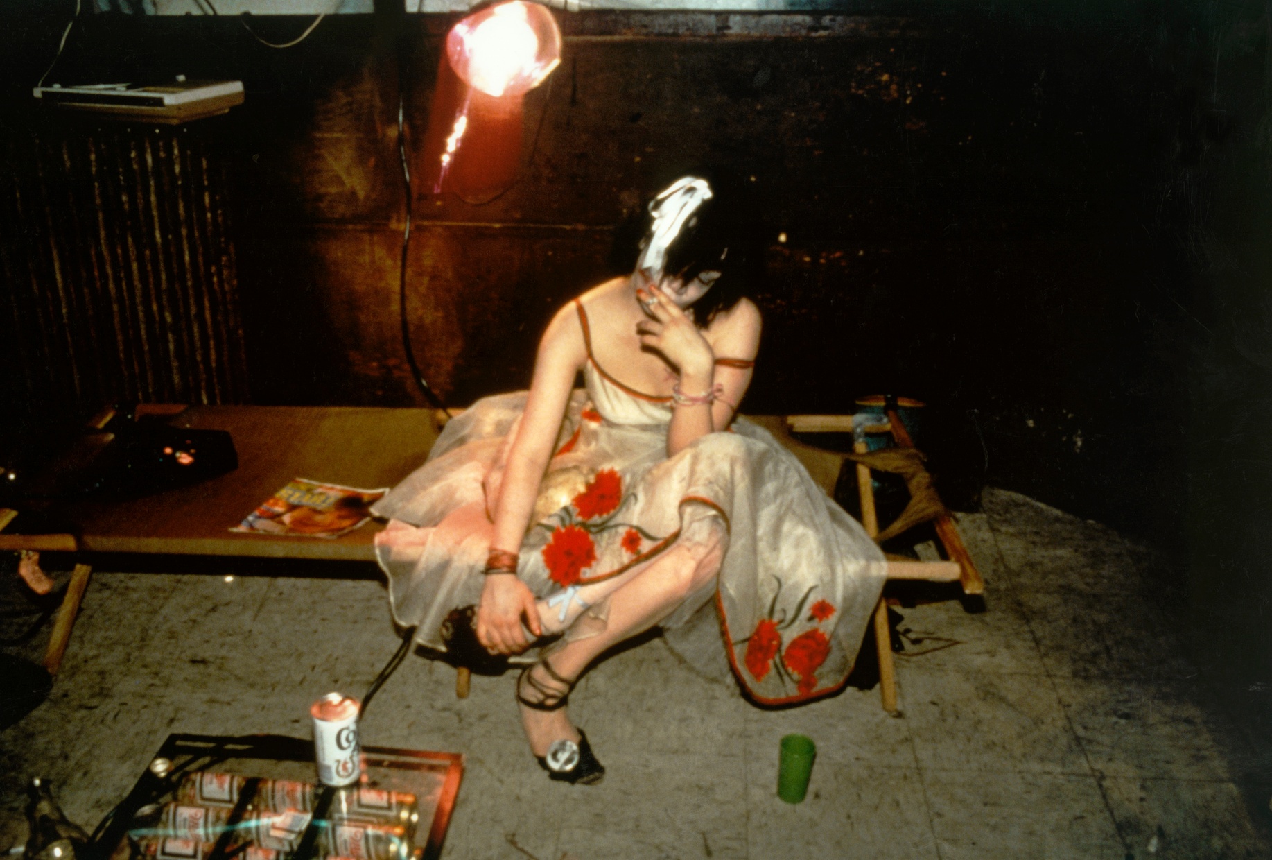 A photograph by Nan Goldin titled "Trixie on the cot, New York City 1979" and features a figure wearing a dress sitting on a coffee table with legs crossed, smoking a cigarette