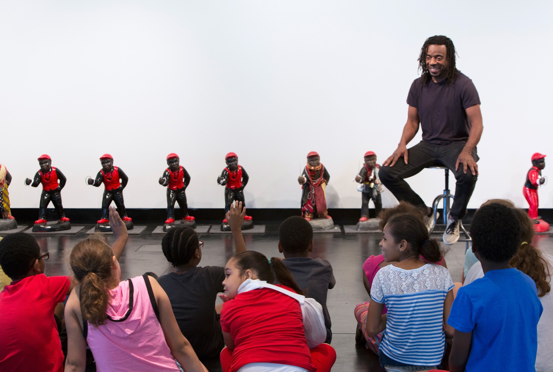 A middle-aged, black man sits, smiling, facing a group of children sitting on a dark floor. A row of painted lawn jockeys are displayed behind the man against a white wall.