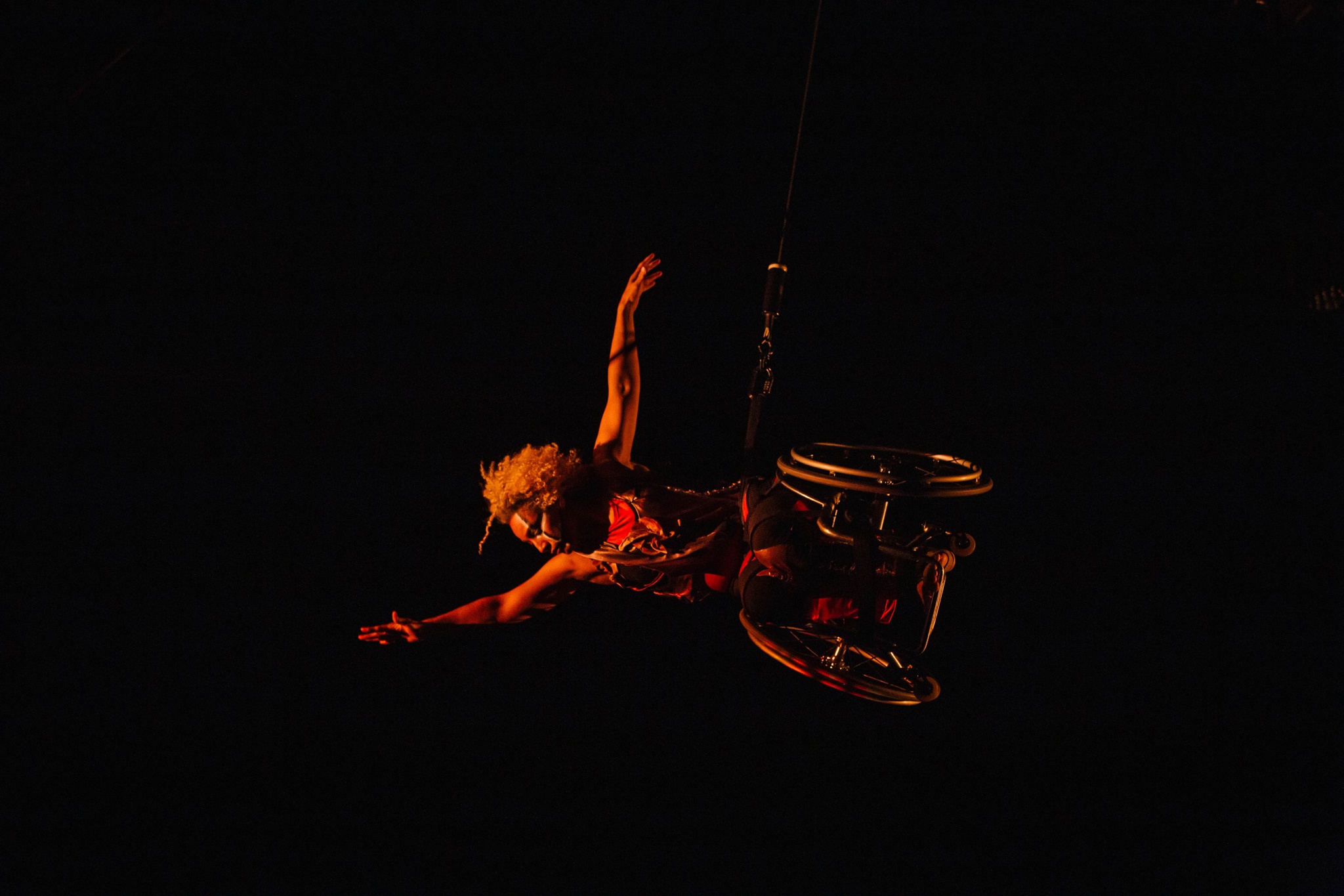 Alice Sheppard flies in her wheelchair, body parallel to the stage as she reaches one arm high and extends the other to the side. She dances in a glowing red shadow. She is a multiracial Black woman with coffee colored skin and short curly hair; she wears a red and gold bodysuit, black and white lines adorn her face.