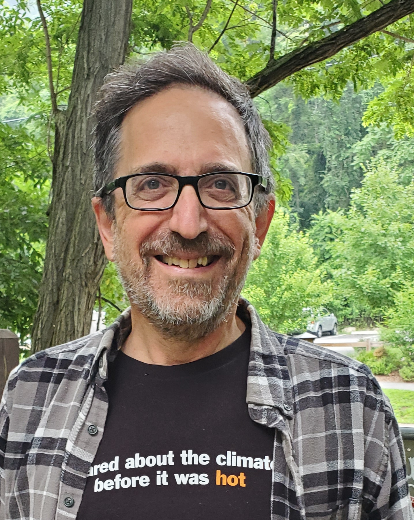 A man with short, salt-and-pepper hair and wearing glasses smiles broadly. His shirt has partially legible words that say "about the climate before it was hot." In the background are green, leafy trees. 