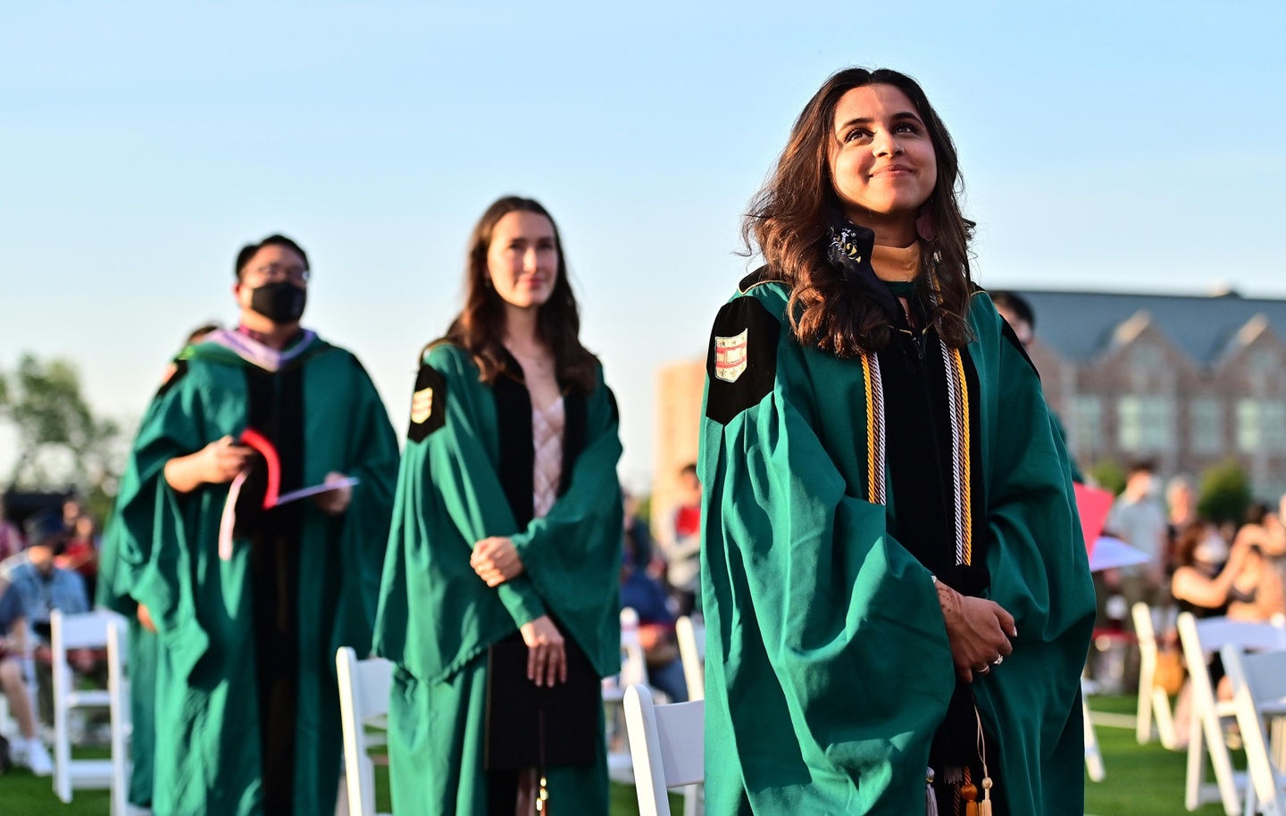 Three students standing in green Commencement gowns outdoors on a blue sky day.