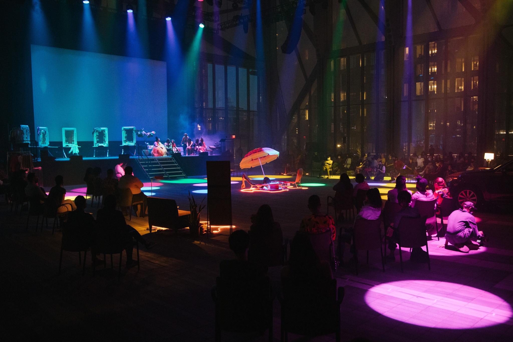 A performance space lit in blue and purple spotlights. The lights illuminate vignettes including a beach chair and umbrella as well as a stage with makeup vanities lining it along the back.