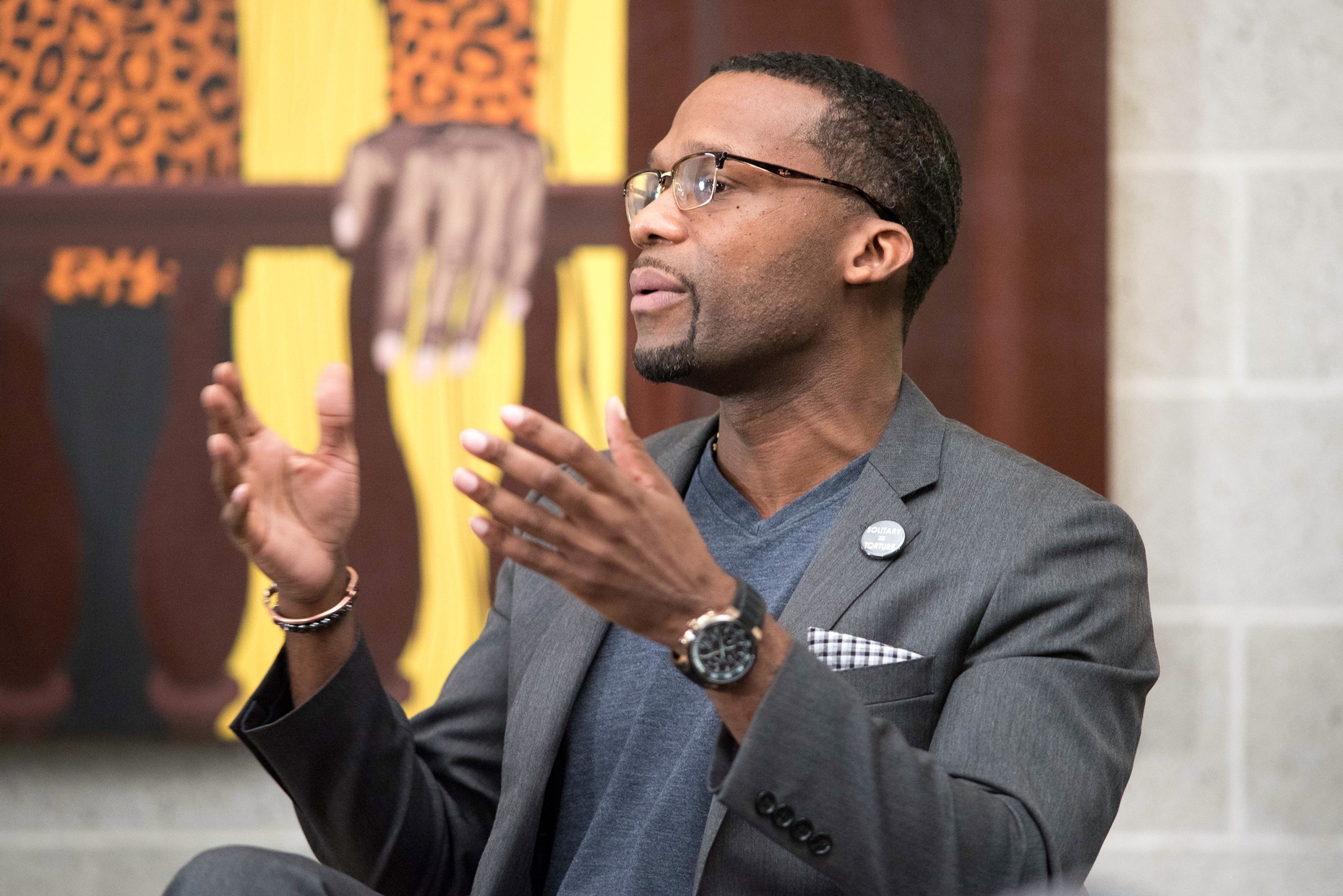 A middle-aged, black man talking with his hands raised in front of a colorful painting.