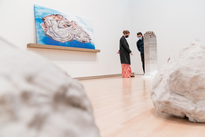 Two people stand and investigate a 4ft tall stone obelisk that transitions from smooth and white at the base to grey and rocky at the top. Blurry in the foreground are some grey boulders, and on the wall is a topographical painting with a blue background.