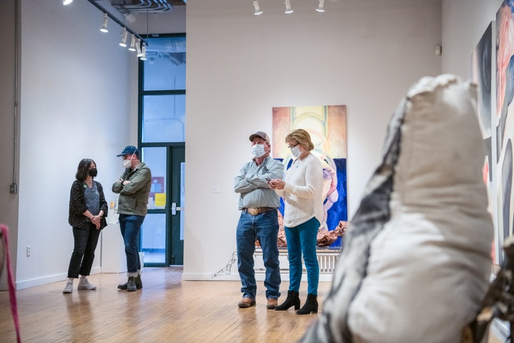 People stand in a gallery space with paintings and soft sculptures visible.