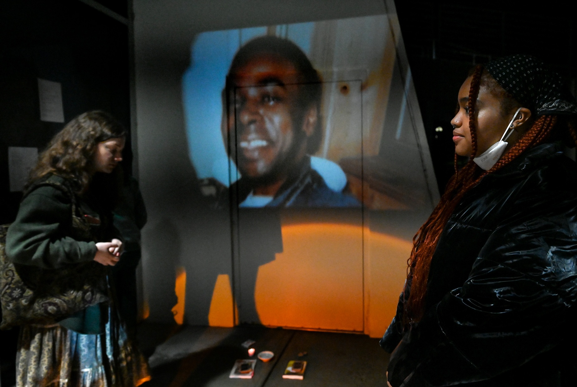 Two people stand in front of a photo of a man projected onto the side of a building.