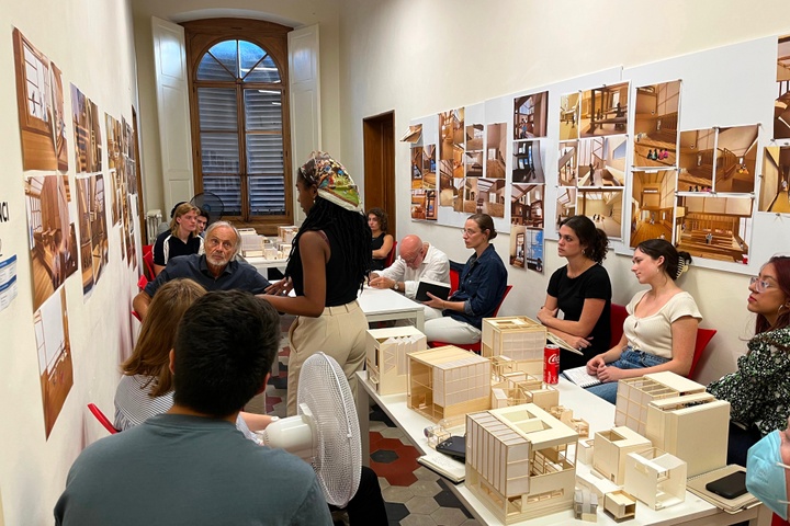 Students gather around a table with white architectural models. one student is presenting their project.