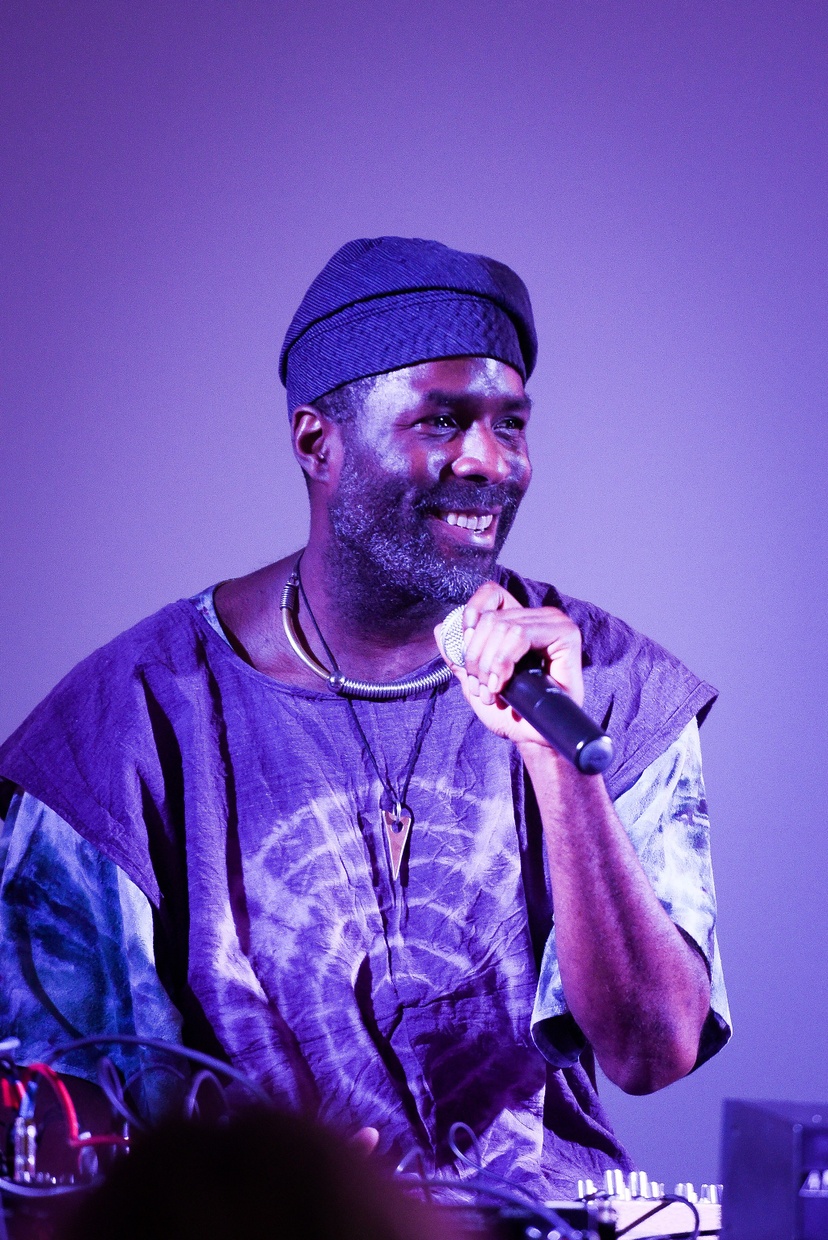 Photo of a man smiling holding a microphone to his mouth in purple lighting.