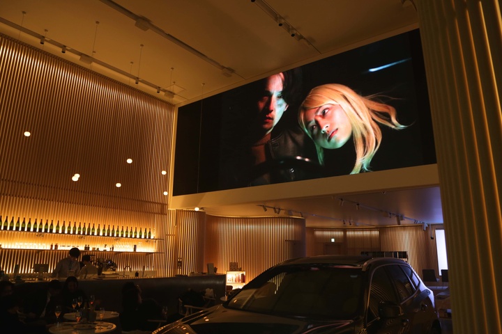 Installation view featuring a video on a wide, high screen showing two figures in the dark. Installation is set in a sleek-looking, dimly lit bar; a car is parked under the video screen next to a couple of round tables with drinks.