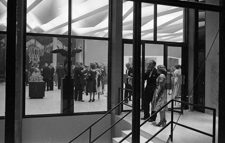 Black and white photo of the foyer of a modern-looking building with a pleated ceiling. People in fancy dress are gathered inside, one person stands in the foyer and smokes a cigarette.
