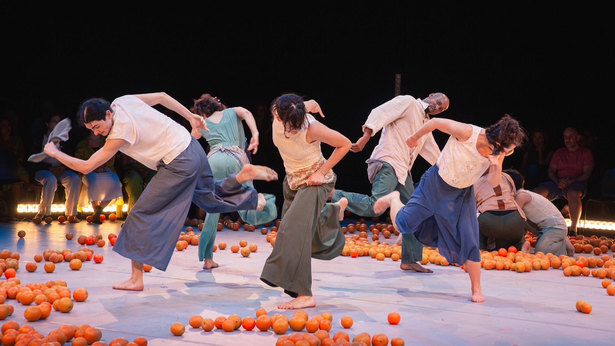 Five dancers from Yaa Samar Dance Theatre perform in a circle. They each bend over with arms out as if running in place. Around them are strewn brightly colored oranges.