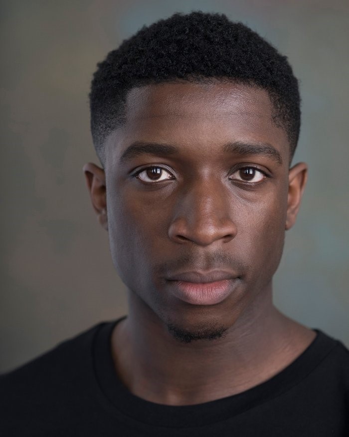 A headshot of actor Caleb Obediah, who looks directly at us with casual intensity. Caleb has dark brown skin, short hair, and wears a black crew neck tshirt. 