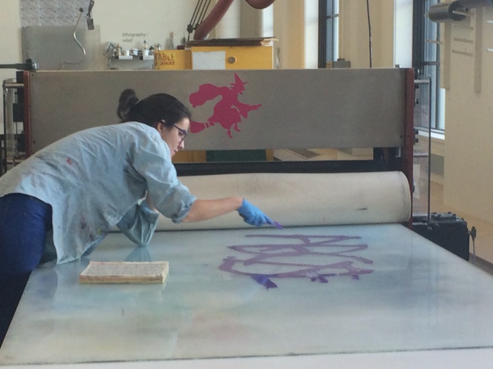 A student bent over a glass panel with a paintbrush working on a print