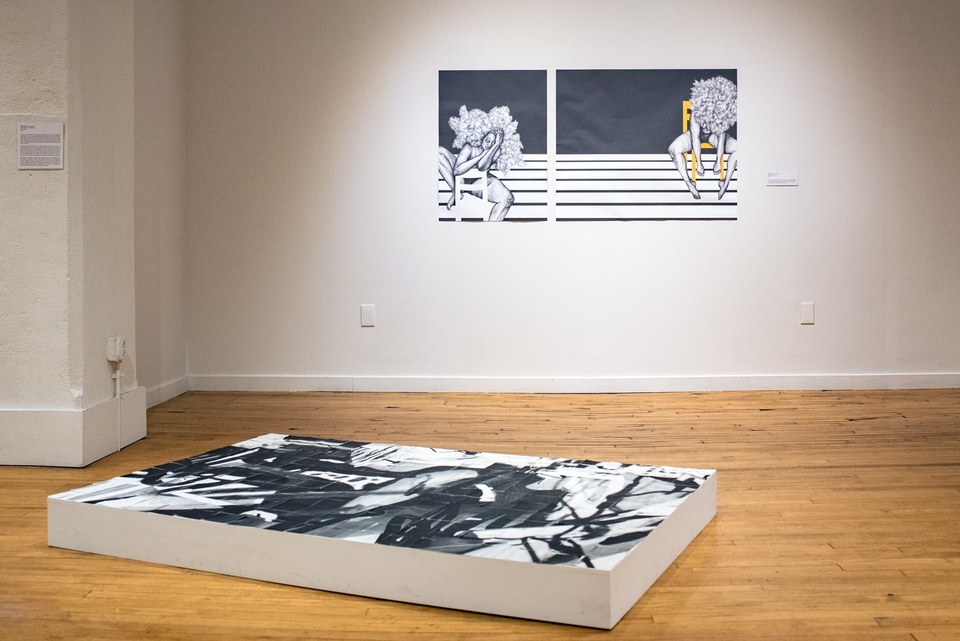 Two artworks in a gallery. On the floor is a white rectangular piece, about 6" high, with a black and white image cut up and jigsawed back together on top. On the wall are two black and white drawings of nude women with huge clouds of untamed hair sitting on chairs in an empty space. 