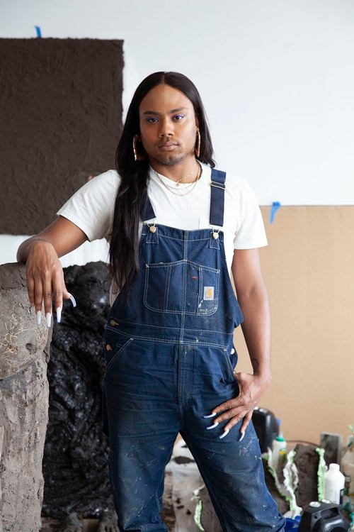 A portrait of Kiyan Williams standing in what looks like an artist studio space. Behind them large pieces of black and brown paper are taped to the wall. Kiyan wears overalls, has long white acrylic nails, and long black hair. 