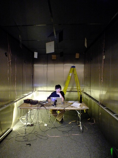 A light-skinned man sits at a table in a dark elevator working on a laptop with a yellow ladder behind him. Cords litter the floor and run up the sides of the elevator and panels hang from the ceiling.