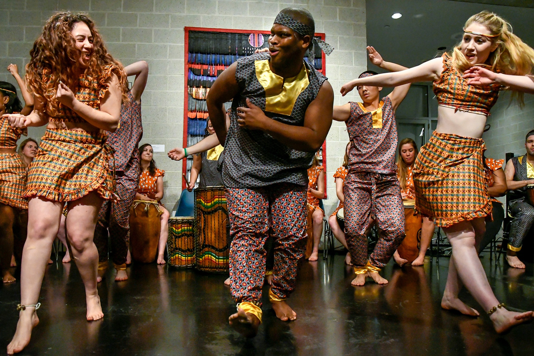 Three young people, one male and two females, wearing colorful, patterned clothing dance with one foot out and arms raised up to their shoulders.
