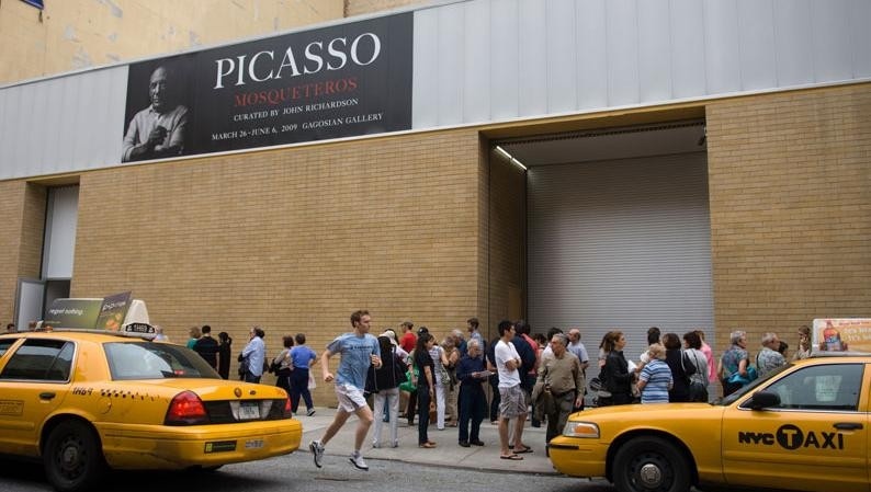 Busy street scene, with two yellow cabs and throngs of people outside a building with a banner advertising the exhibition Picasso: Mosqueteros.