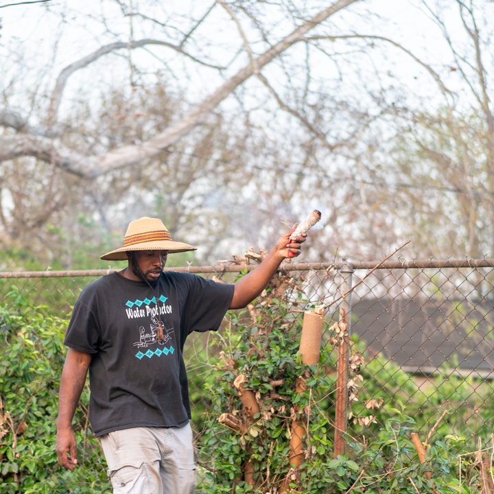 Photo shows a man wearing a hat and a black teeshirt carrying an object in his hand as he walks along a chainlink fence outdoors.