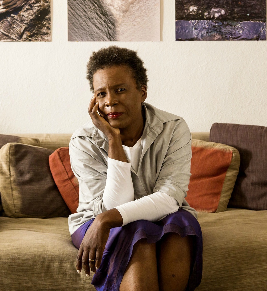 Claudia Rankine sitting on a brown couch with artwork behind her and head propped on hand.