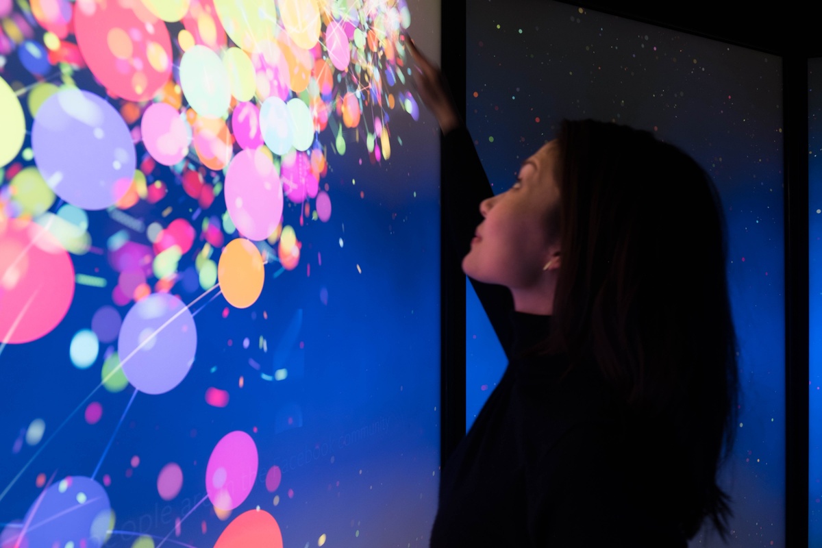 Young woman interacting with screen from the side, touching her hand to the screen which generates a trail of visual graphics