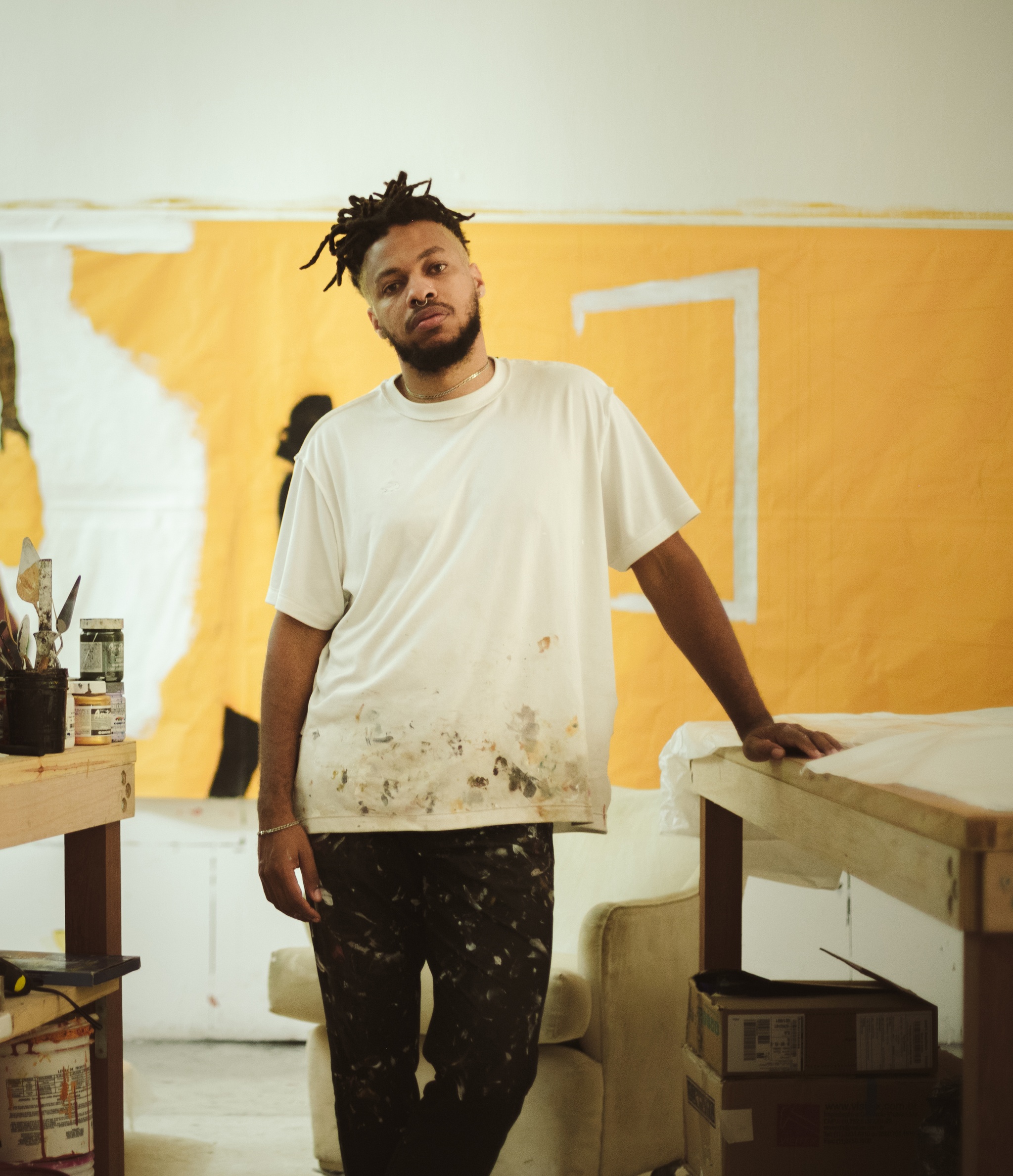 A portrait of artist Maxwell Alexandre standing in his studio with a yellow brown painting on the wall behind him. Maxwell is a Black Brazilian man with a beard. He wears a white t-shirt that is dirty with paint at the bottom hem.