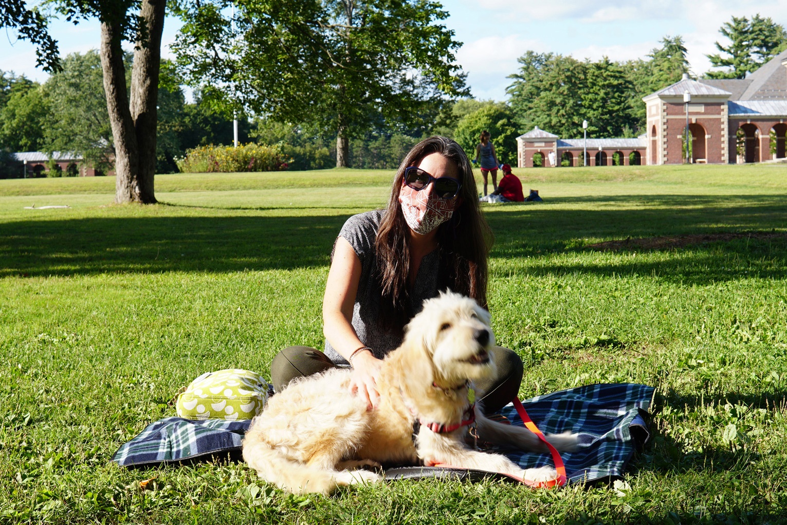 A light-skinned woman with dark hair wearing sunglasses and a red mask with white leaves and lines printed on it petting a cream dog sitting on a blanket on grass.