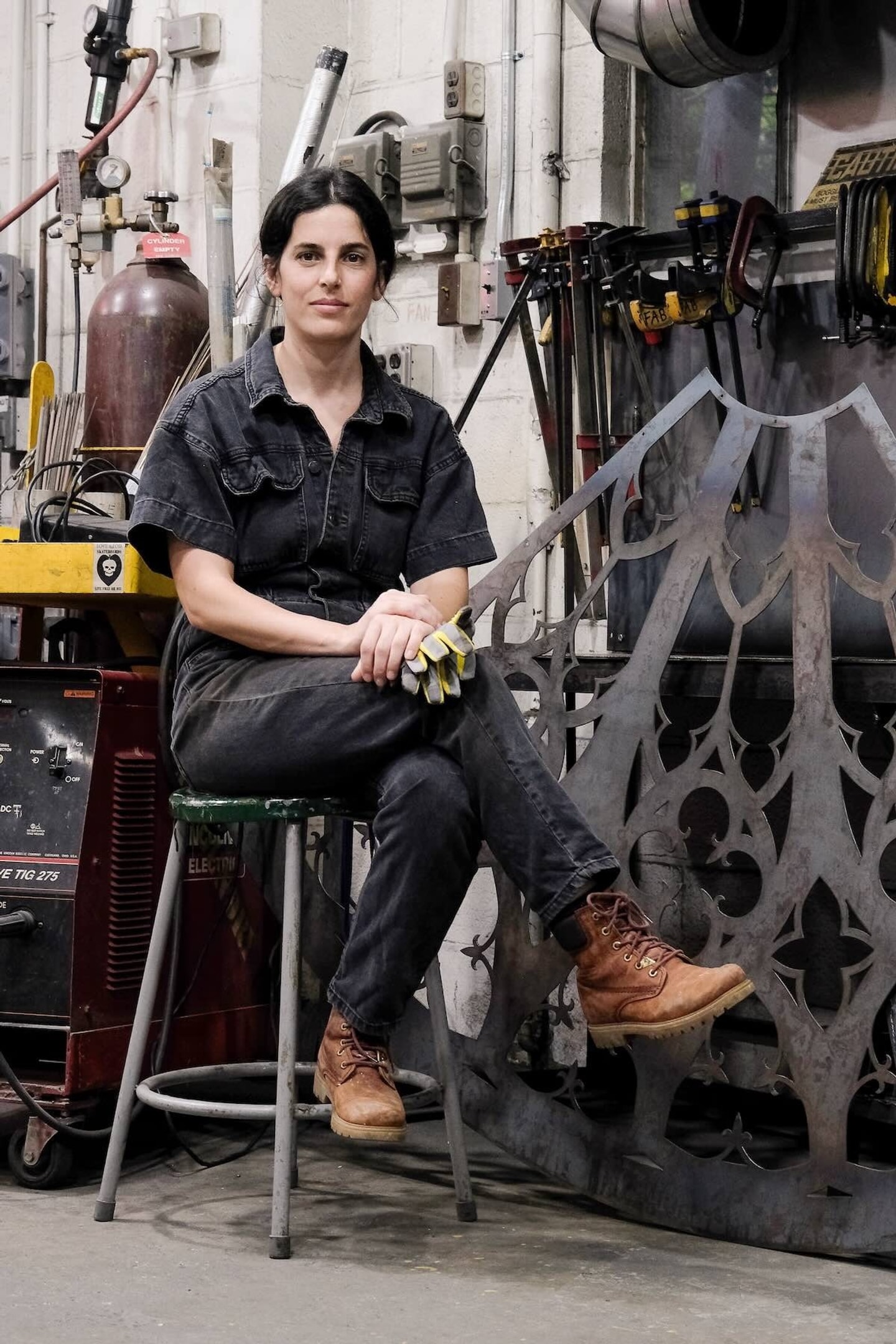 artist sitting on a stool next to machinery in coveralls with work boots and her hair pulled back