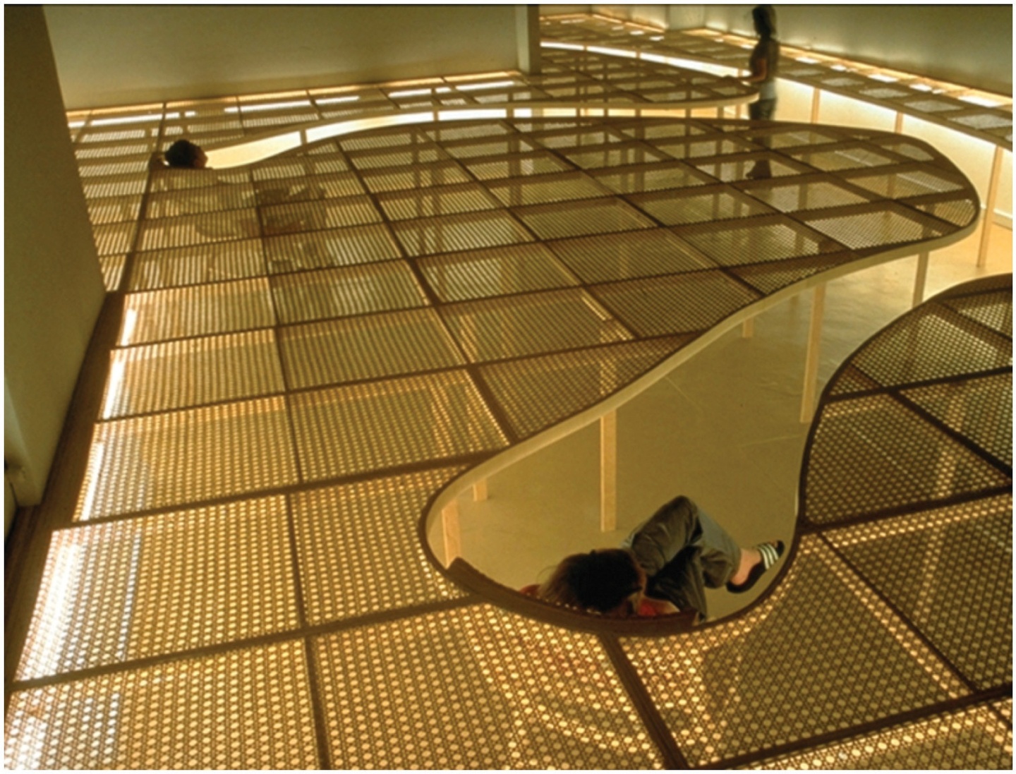 A dimly lit space, fluorescent lights illuminating the edge of a raised level of see-through architectural forms: two people sit in chairs below, as if sunken into the ground, so that they can see above the partitions into the vast space. Another person stands in the background.