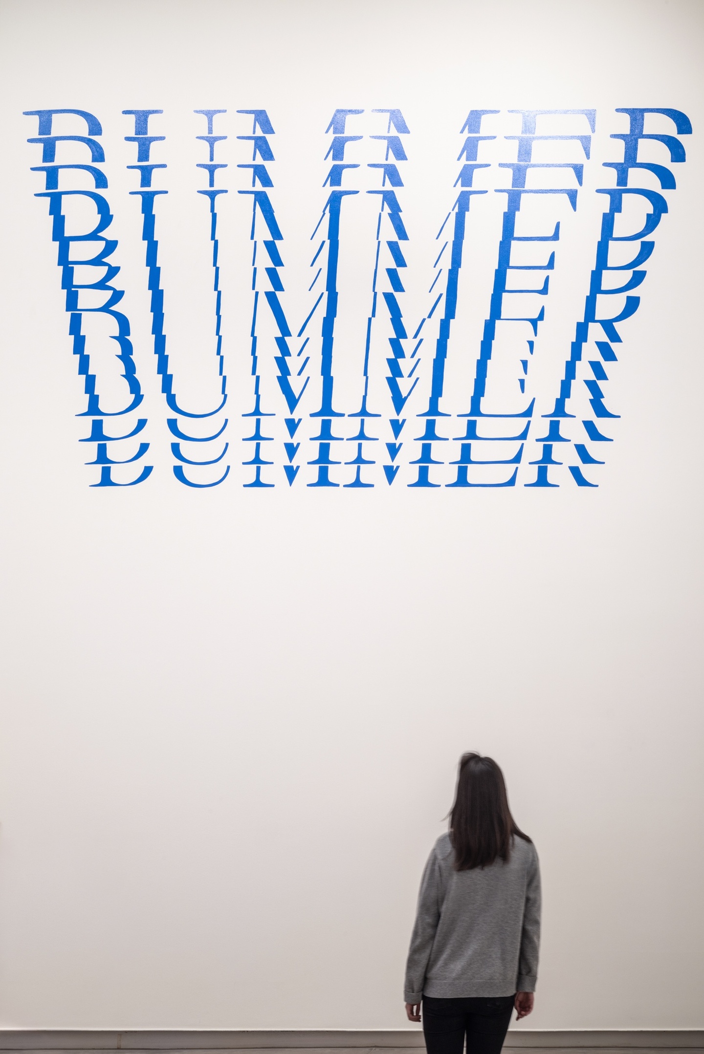 Large blue text on the gallery wall with the word BUMMER repeated in horizontal cutouts