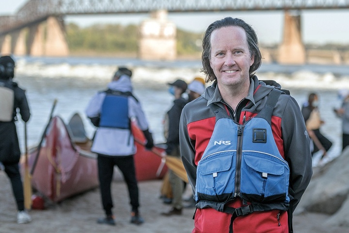 Faculty member Derek Hoeferlin stands, with a life jacket on, in the foreground, with his students preparing to canoe behind him. The river and a bridge are in the background.
