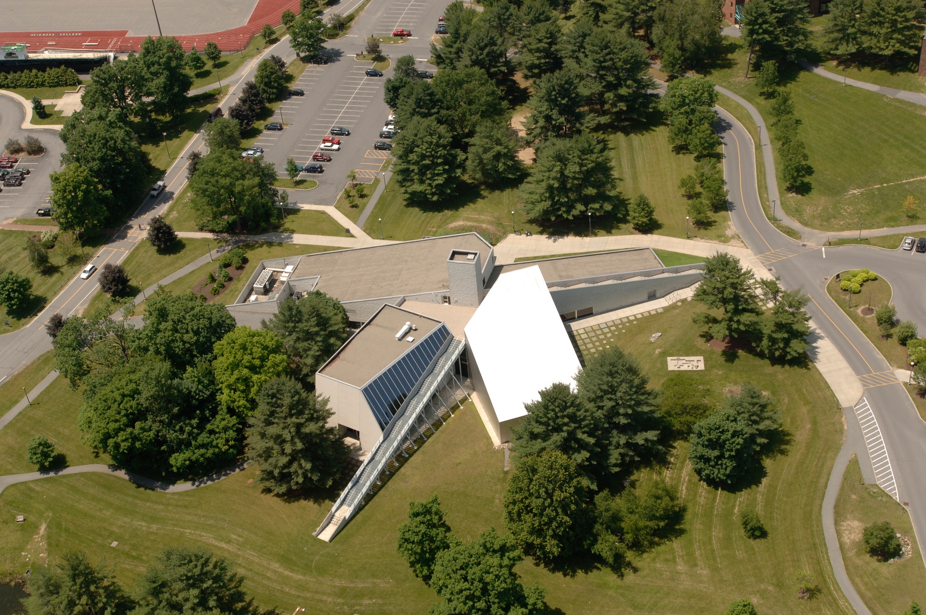 An aerial photograph of a white and gray, angular building that juts out in four different directions surrounded by grass and trees.