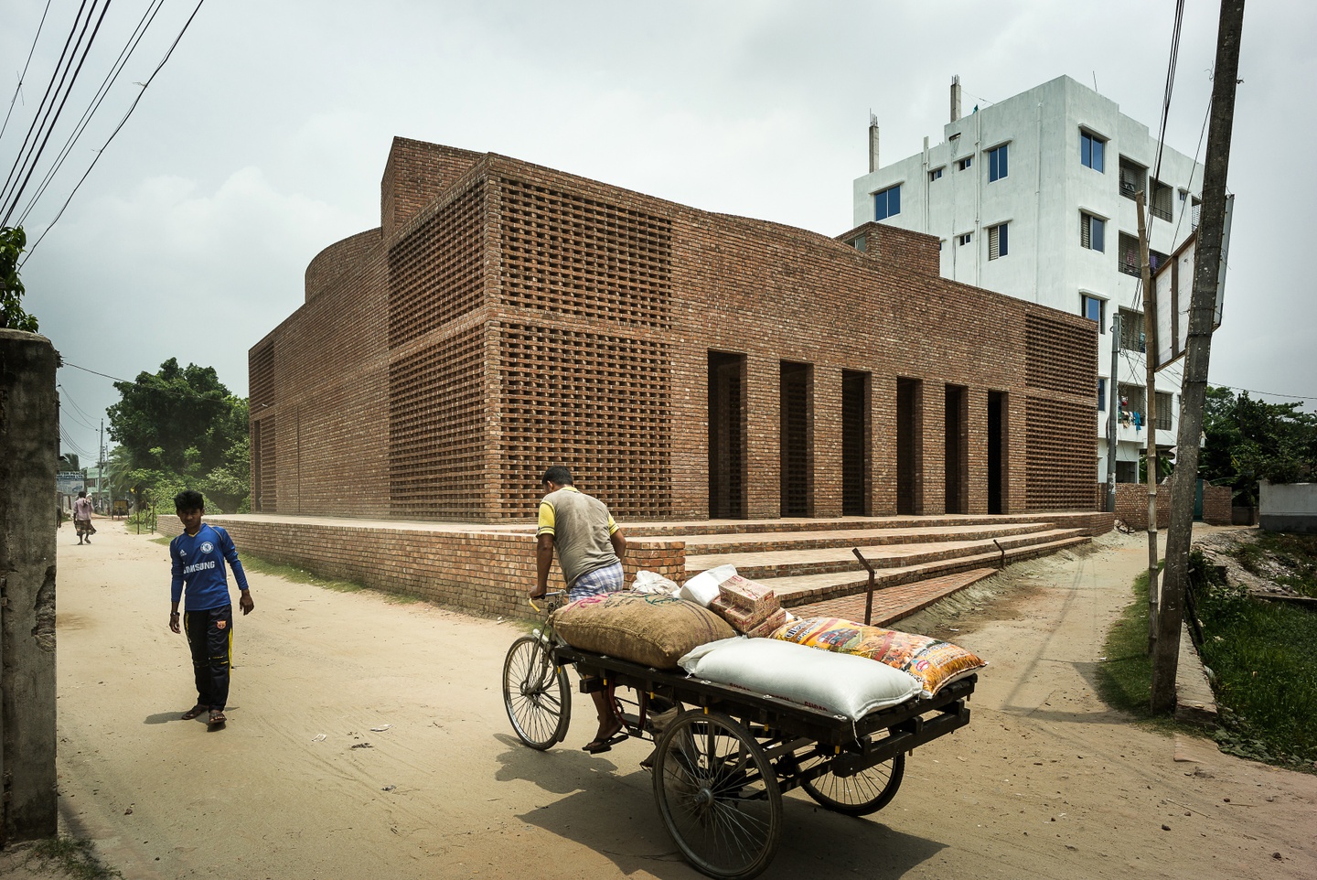 Photo of a red-brick, flat-roofed mosque with several doorway entries; a person on a bike with a cart is in the foreground.