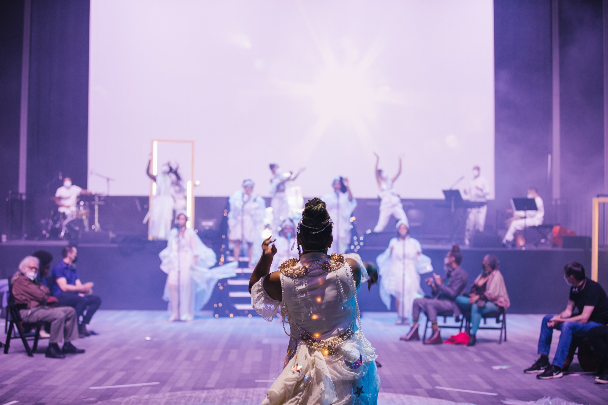 A performer dressed in white with her back to the camera faces a line of performers on a stage in the background. The performance space is lit with a bright pale purple light.