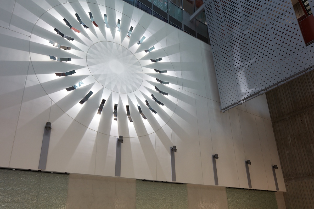 Photograph looking upward of circular metal sculpture on wall with white light radiating outward