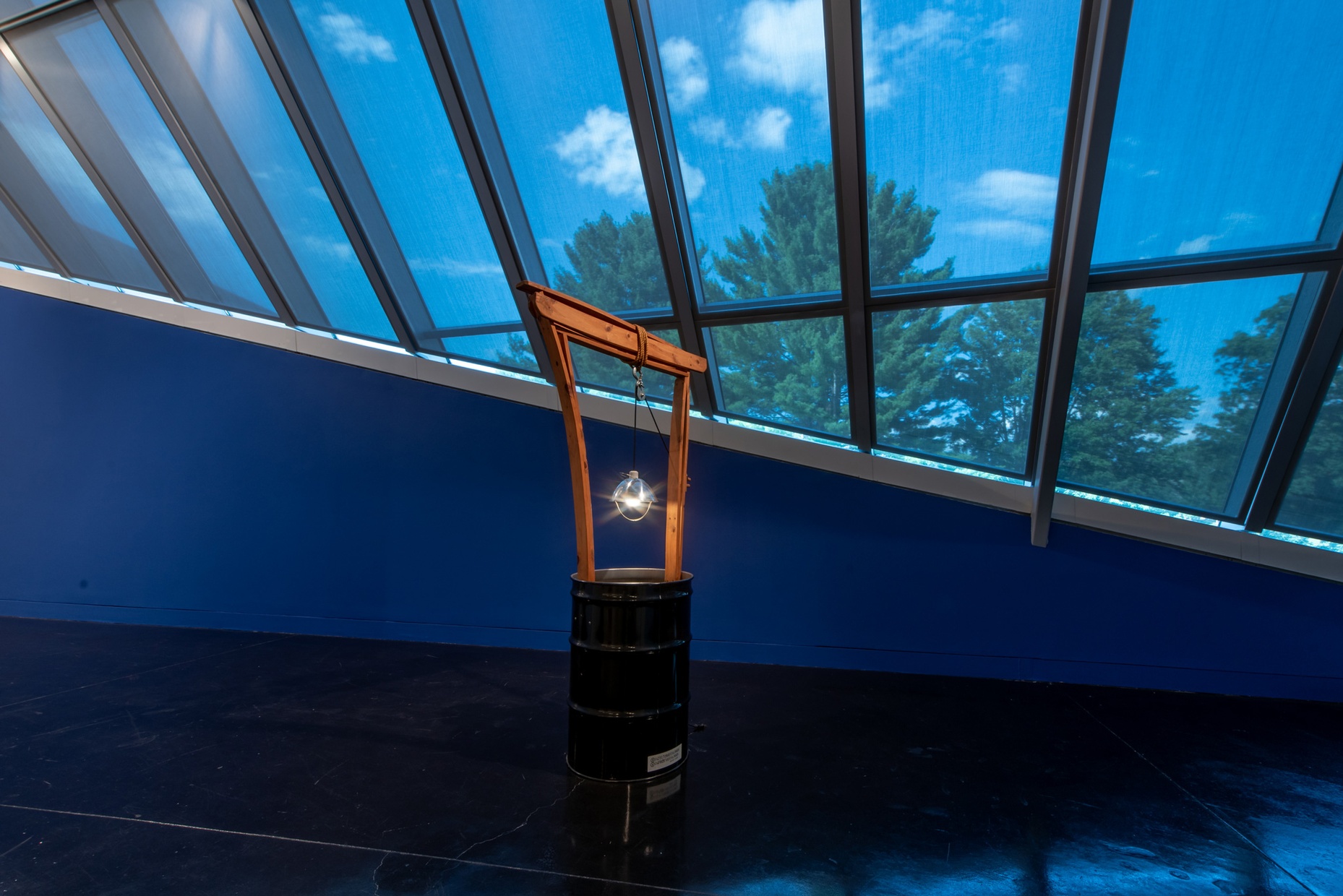 Against a dark blue wall below a large window sits a well-like structure made of a black steel drum out of which protrudes a light hanging from a wooden arch.