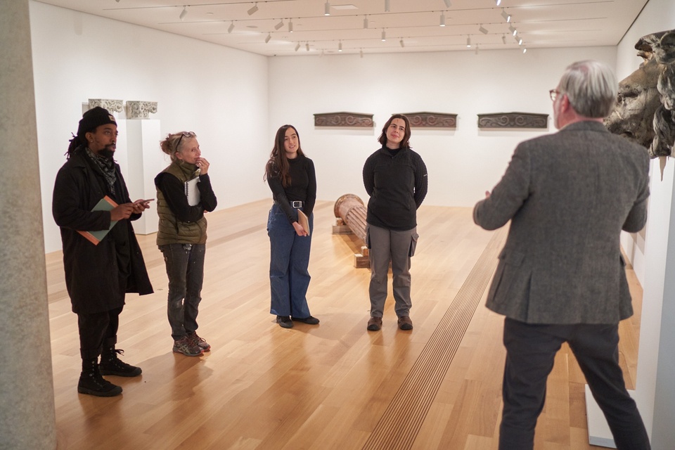 Students stand in a gallery of architectural objects at the Pulitzer as a curator talks.