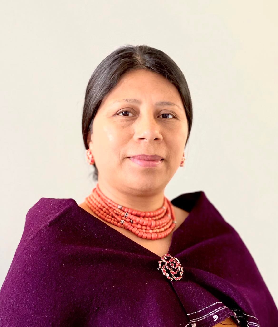 A portrait of Mirian Masaquiza Jerez, an Indigenous Kichwa woman from Ecuador. Her hair is parted in the middle and pulled back smoothly behind her head. She wears a purple cape attached over her chest by a brooch along with a long beaded necklace wrapped in many strands around her neck.