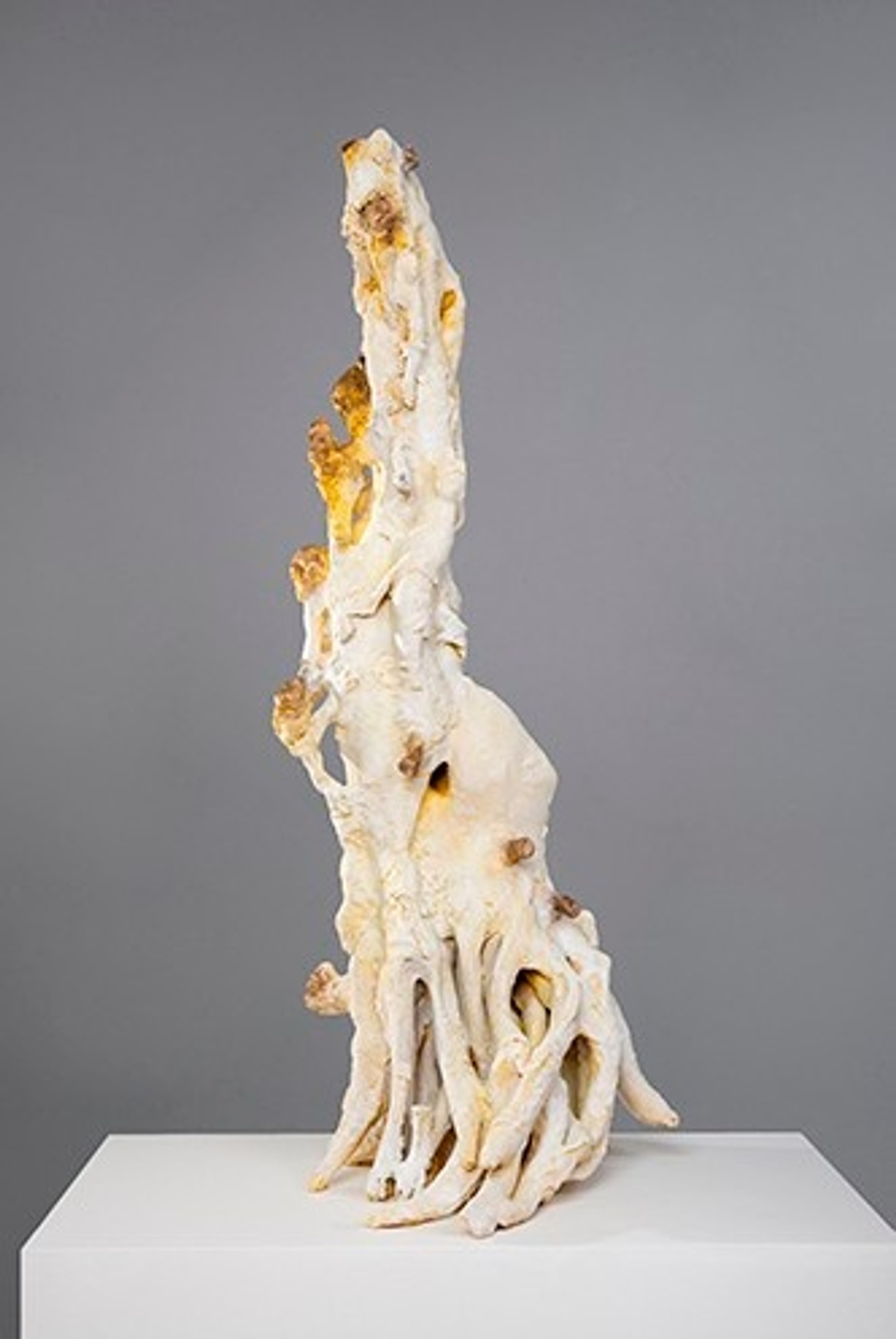 An abstract, tall ceramic sculpture by Arny Nadler featuring pale, intertwining root-like tendrils and a tapering vertical spire with amber-yellow accents, set on a white pedestal against a grey background.