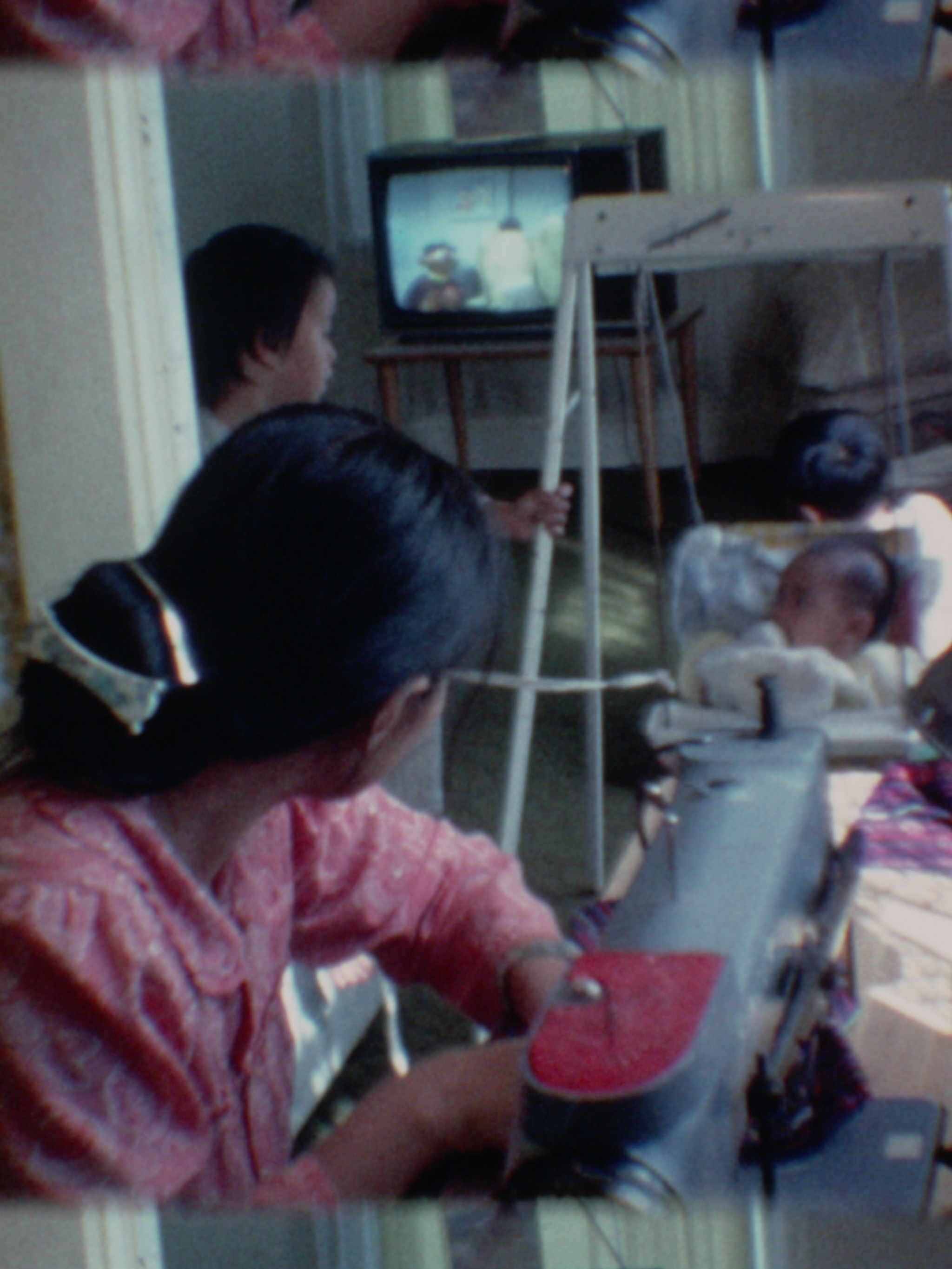A Vietnamese woman sits at her sewing machine, facing away from the camera, her smooth black hair gathered in a low bun, fastened with a gold clip and white, bow-shaped barrette. The woman’s face is turned toward the living room toward three children: a baby in a swing, pulled by a rope in the hand of a young boy in profile, and the back of a young boy’s head. On a 13 inch box television,  which is perched on a wooden table, Burt and Ernie from Sesame Street are on the screen.